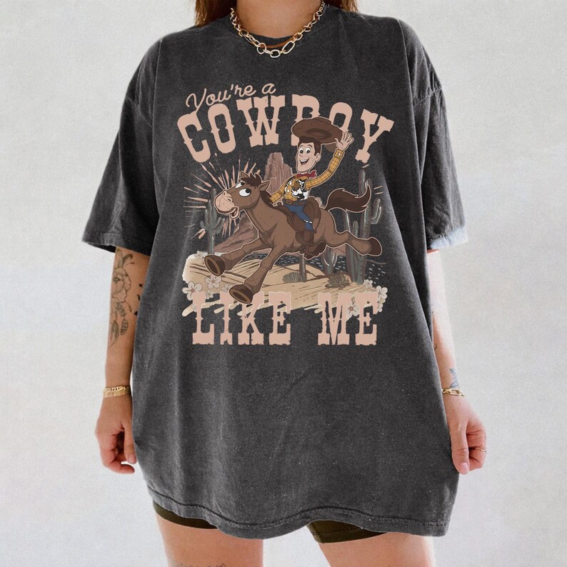 May include: A charcoal grey short-sleeve t-shirt featuring a graphic of a cartoon cowboy riding a horse. The text "You're a COWBOY LIKE ME" is printed in a light beige font above and below the illustration, which includes cacti and desert scenery.