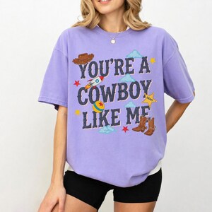 May include: A light purple short-sleeve t-shirt featuring the text "YOU'RE A COWBOY LIKE ME" in dark blue letters. The design includes a brown cowboy hat, a rocket, a colorful ball, a yellow sheriff star, and brown cowboy boots with clouds.