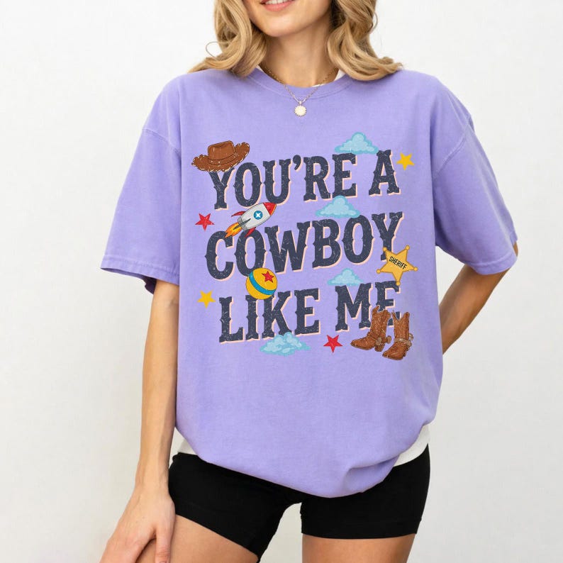 May include: A light purple short-sleeve t-shirt featuring the text "YOU'RE A COWBOY LIKE ME" in dark blue letters. The design includes a brown cowboy hat, a rocket, a colorful ball, a yellow sheriff star, and brown cowboy boots with clouds.