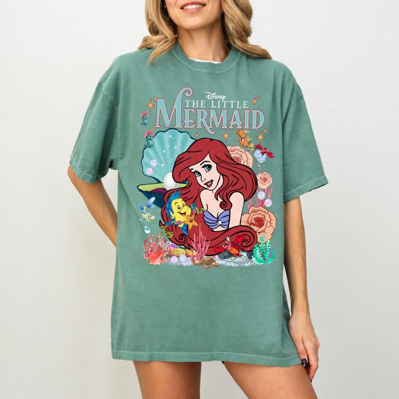 Op de afbeelding: Groen T-shirt met een afbeelding van Ariel uit Disney's De Kleine Zeemeermin. Het ontwerp bevat Ariel, Botje, Sebastiaan en de woorden "The Little Mermaid". Het shirt heeft een ronde hals en korte mouwen.