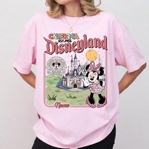 May include: Pink t-shirt featuring a Disneyland graphic. The graphic includes the words "California Disneyland EST. 1955", a cartoon of Minnie Mouse, a castle, and a Ferris wheel. The shirt has short sleeves and a crew neck.