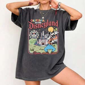 May include: Dark gray t-shirt featuring a colorful graphic of Disneyland with Goofy. The design includes a Ferris wheel, castle, and the text "California Disneyland Est. 1955". The shirt has a vintage, worn look.