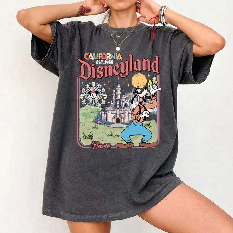 May include: Dark gray t-shirt featuring a colorful graphic of Disneyland with Goofy. The design includes a Ferris wheel, castle, and the text "California Disneyland Est. 1955". The shirt has a vintage, worn look.