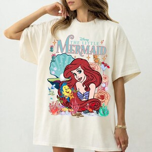 Op de afbeelding: Cr&egrave;mekleurig T-shirt met een kleurrijke afbeelding van Ariel uit Disney's De Kleine Zeemeermin. Het ontwerp bevat Ariel, Botje en andere zeedieren, met de filmtitel in een gestileerd lettertype. Het shirt heeft korte mouwen.