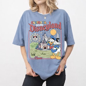 May include: A light blue t-shirt featuring a Disneyland graphic. The graphic includes the words "California Disneyland EST.1955" and a cartoon image of Donald Duck waving in front of a castle and a Ferris wheel.