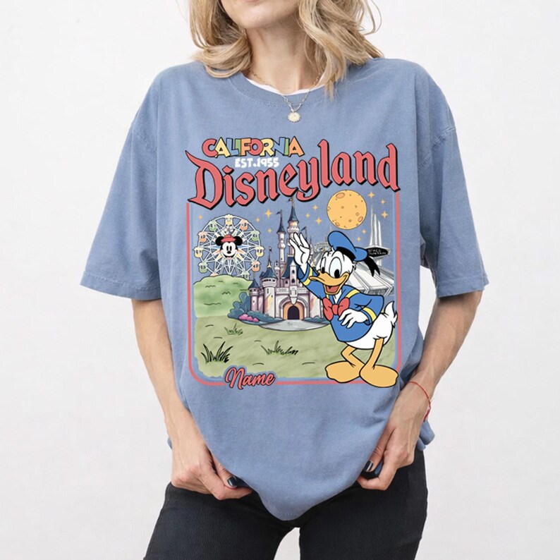 May include: A light blue t-shirt featuring a Disneyland graphic. The graphic includes the words "California Disneyland EST.1955" and a cartoon image of Donald Duck waving in front of a castle and a Ferris wheel.