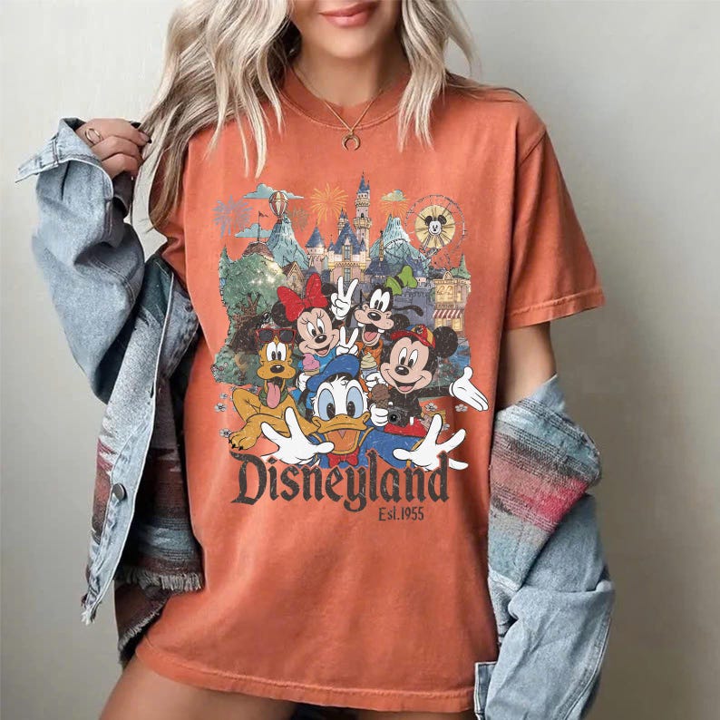 May include: A burnt orange crew neck t-shirt featuring a graphic print of Mickey Mouse, Minnie Mouse, Donald Duck, Goofy, and Pluto in front of a castle. The text Disneyland Est. 1955 is printed below the characters in a dark, stylized font.