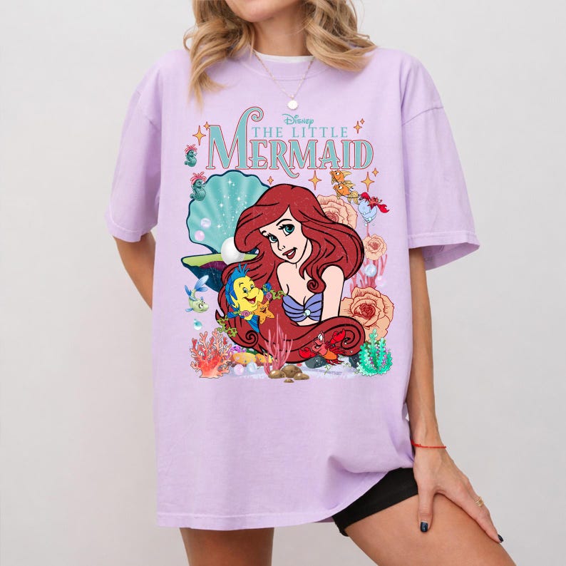 Op de afbeelding: Een lila T-shirt met een afbeelding van Ariel uit Disney's De Kleine Zeemeermin. Het ontwerp bevat Ariel, Botje, Sebastiaan en andere zeedieren, met de filmtitel in een turquoise lettertype.