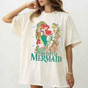 Op de afbeelding: Cr&egrave;mekleurig T-shirt met een kleurrijke afbeelding van Ariel uit De Kleine Zeemeermin. Het ontwerp bevat Ariel, Botje en bloemelementen, met de woorden "The Little Mermaid" in teal.