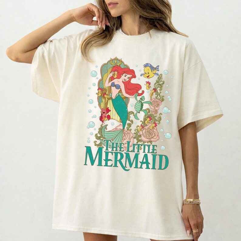 Op de afbeelding: Cr&egrave;mekleurig T-shirt met een kleurrijke afbeelding van Ariel uit De Kleine Zeemeermin. Het ontwerp bevat Ariel, Botje en bloemelementen, met de woorden "The Little Mermaid" in teal.