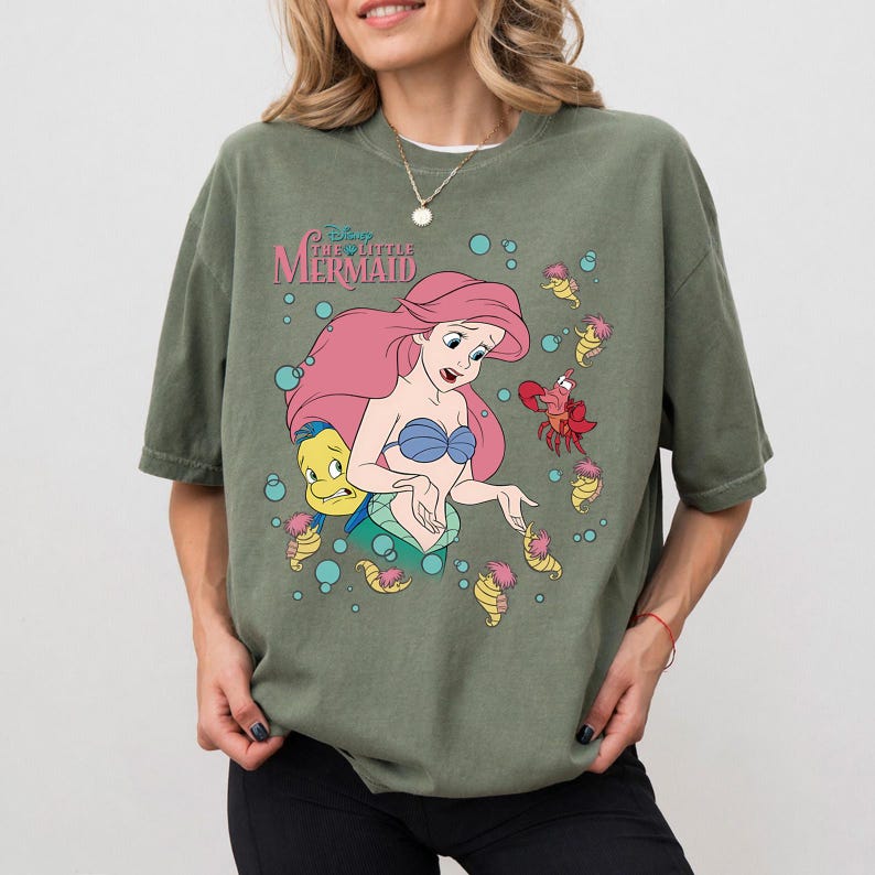 May include: An olive green t-shirt featuring a graphic print of Ariel from The Little Mermaid with long pink hair, a blue seashell top, and a green tail. The design includes Flounder, Sebastian, and small sea creatures surrounded by blue bubbles.