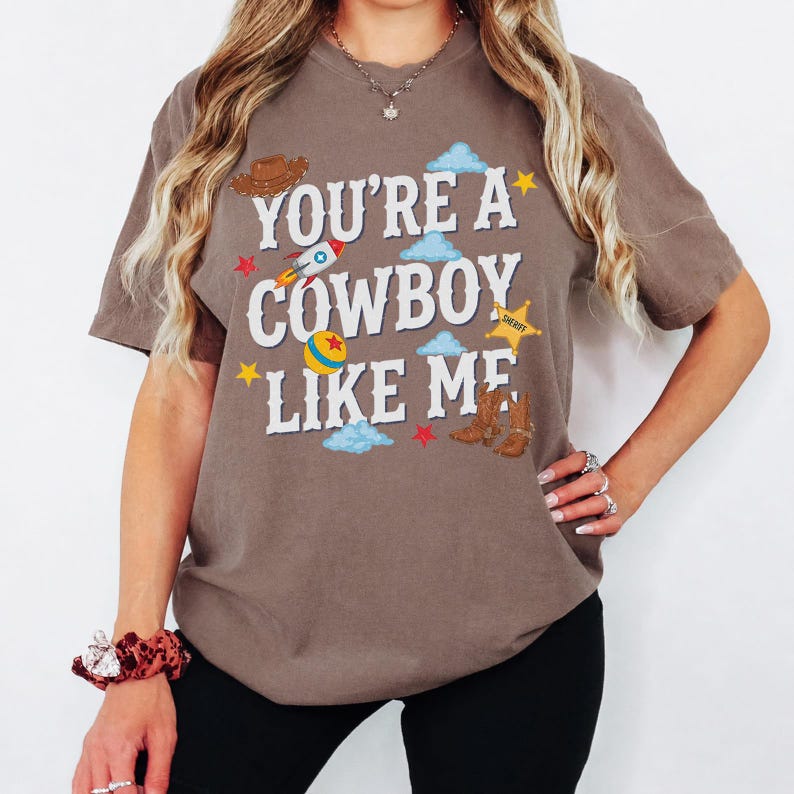 May include: A brown short-sleeve t-shirt featuring the white text "YOU'RE A COWBOY LIKE ME" surrounded by graphic illustrations of a brown cowboy hat, a rocket ship, a yellow ball, a yellow sheriff star, brown cowboy boots, and blue clouds.