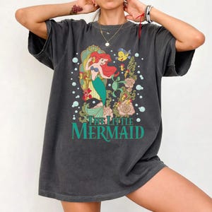 Op de afbeelding: Donkergrijs t-shirt met een afbeelding van Ariel uit De Kleine Zeemeermin. Ariel heeft rood haar en een groene staart, omringd door zeedieren en bloemen. De tekst "THE LITTLE MERMAID" is gedrukt in turquoise.