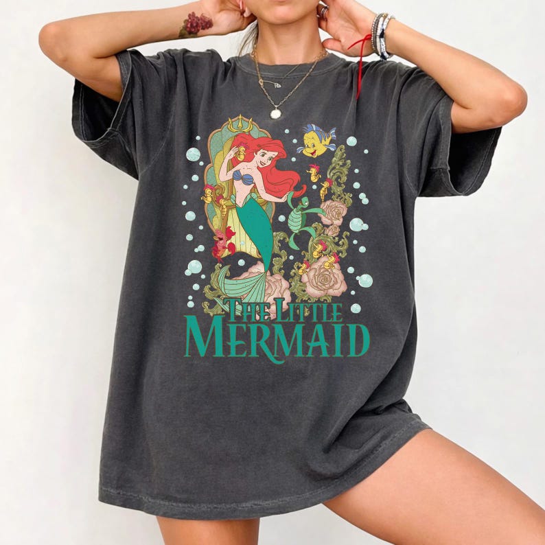 Op de afbeelding: Donkergrijs t-shirt met een afbeelding van Ariel uit De Kleine Zeemeermin. Ariel heeft rood haar en een groene staart, omringd door zeedieren en bloemen. De tekst "THE LITTLE MERMAID" is gedrukt in turquoise.