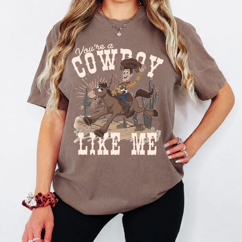 May include: A brown short-sleeve t-shirt featuring a graphic of a cartoon cowboy riding a horse. The text "You're a COWBOY LIKE ME" is printed in a cream-coloured western-style font above and below the illustration, which includes cacti.