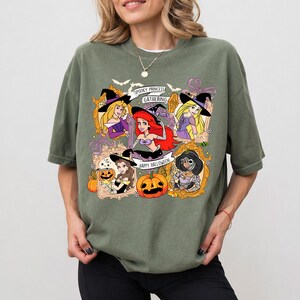 May include: Olive green t-shirt featuring a Halloween-themed graphic. The design includes cartoon princesses in witch hats, pumpkins, and the text "Spooky Princess Gathering" and "Happy Halloween".