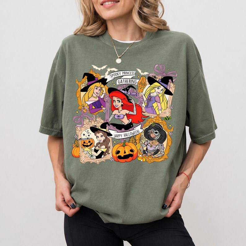 May include: Olive green t-shirt featuring a Halloween-themed graphic. The design includes cartoon princesses in witch hats, pumpkins, and the text "Spooky Princess Gathering" and "Happy Halloween".