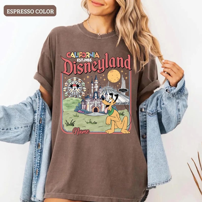 May include: Espresso-colored t-shirt featuring a Disneyland graphic. The design includes the words "California Disneyland EST. 1955" with illustrations of a castle, Pluto, and a Ferris wheel. The shirt has rolled-up sleeves.