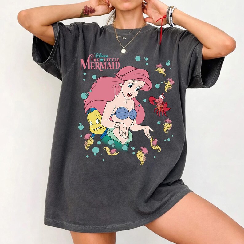 May include: A charcoal grey short-sleeve t-shirt featuring a graphic print of Ariel from The Little Mermaid with Flounder and Sebastian. The design includes teal bubbles and small yellow seahorses surrounding the characters on the front.