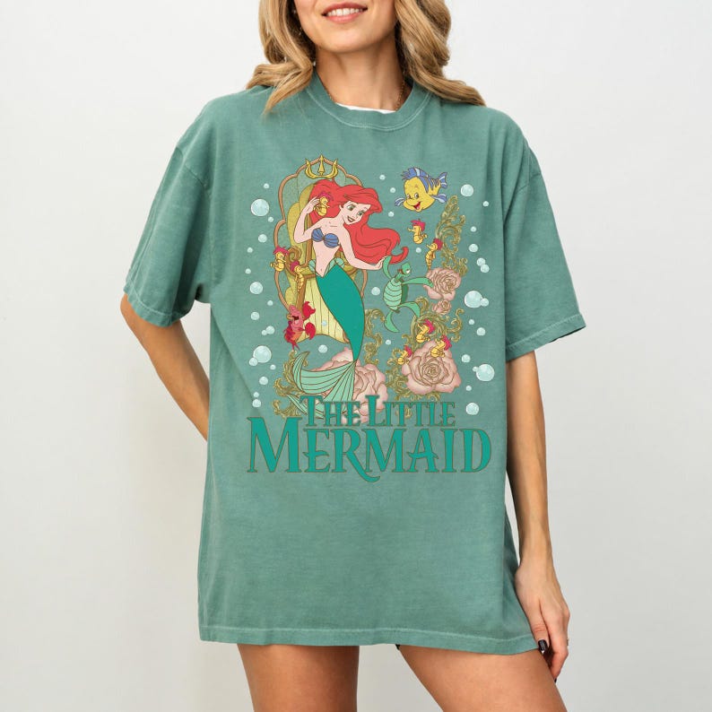 Op de afbeelding: Een zeegroen T-shirt met een afbeelding van Ariel uit De Kleine Zeemeermin. Het ontwerp toont Ariel met rood haar en een groene staart, met de tekst "THE LITTLE MERMAID". Het shirt heeft een losse pasvorm.