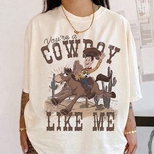 May include: An off-white short-sleeve t-shirt featuring a graphic of a cartoon cowboy riding a brown horse in a desert landscape with cacti. The text reads You're a COWBOY LIKE ME in a brown western-style font above and below the illustration.