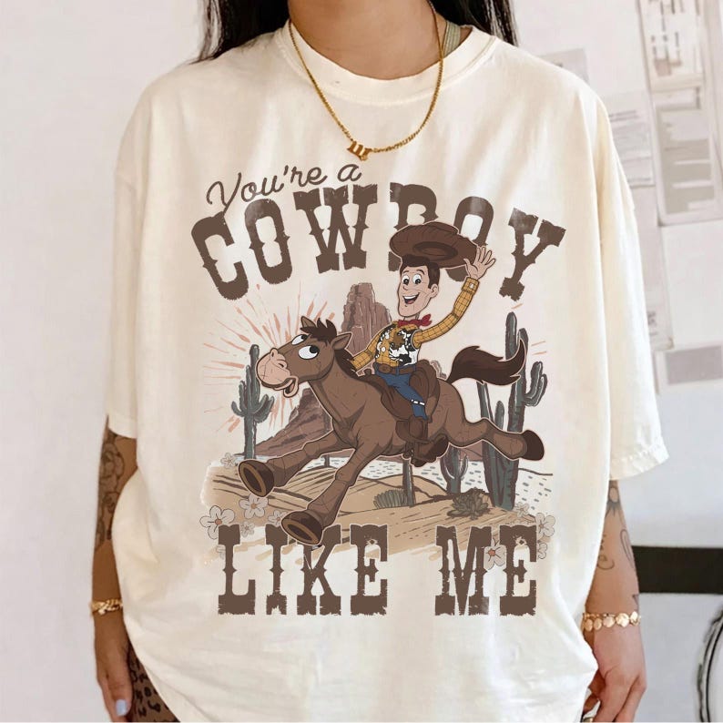 May include: An off-white short-sleeve t-shirt featuring a graphic of a cartoon cowboy riding a brown horse in a desert landscape with cacti. The text reads You're a COWBOY LIKE ME in a brown western-style font above and below the illustration.