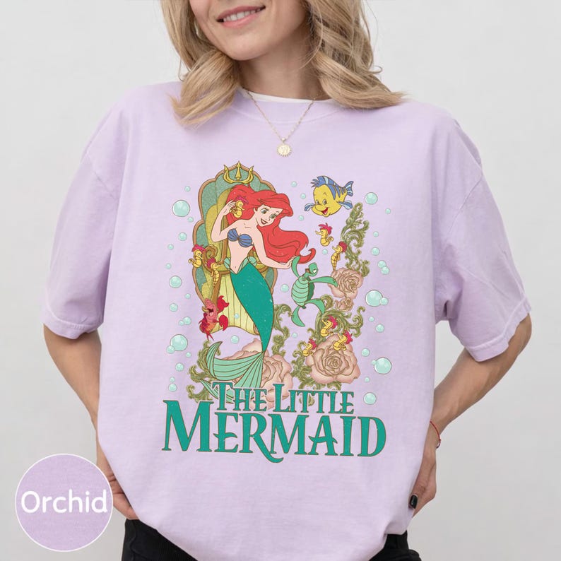 Op de afbeelding: Orchidee-kleurig T-shirt met een afbeelding van Ariel uit De Kleine Zeemeermin. Het ontwerp toont Ariel met rood haar en een groene staart, samen met Botje en de tekst "THE LITTLE MERMAID" in het groen.