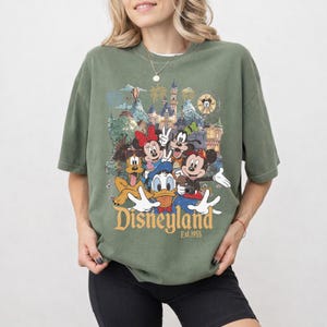 May include: An olive green short-sleeve t-shirt featuring a graphic print of Mickey Mouse, Minnie Mouse, Donald Duck, Goofy, and Pluto in front of a castle. The text "Disneyland Est. 1955" is printed in a gold serif font below the character illustration.