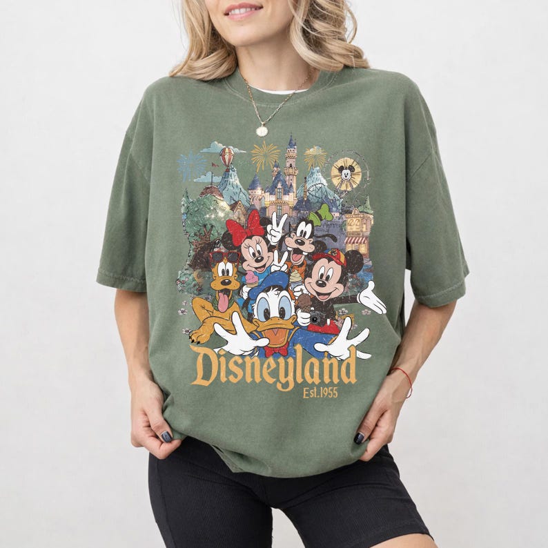 May include: An olive green short-sleeve t-shirt featuring a graphic print of Mickey Mouse, Minnie Mouse, Donald Duck, Goofy, and Pluto in front of a castle. The text "Disneyland Est. 1955" is printed in a gold serif font below the character illustration.