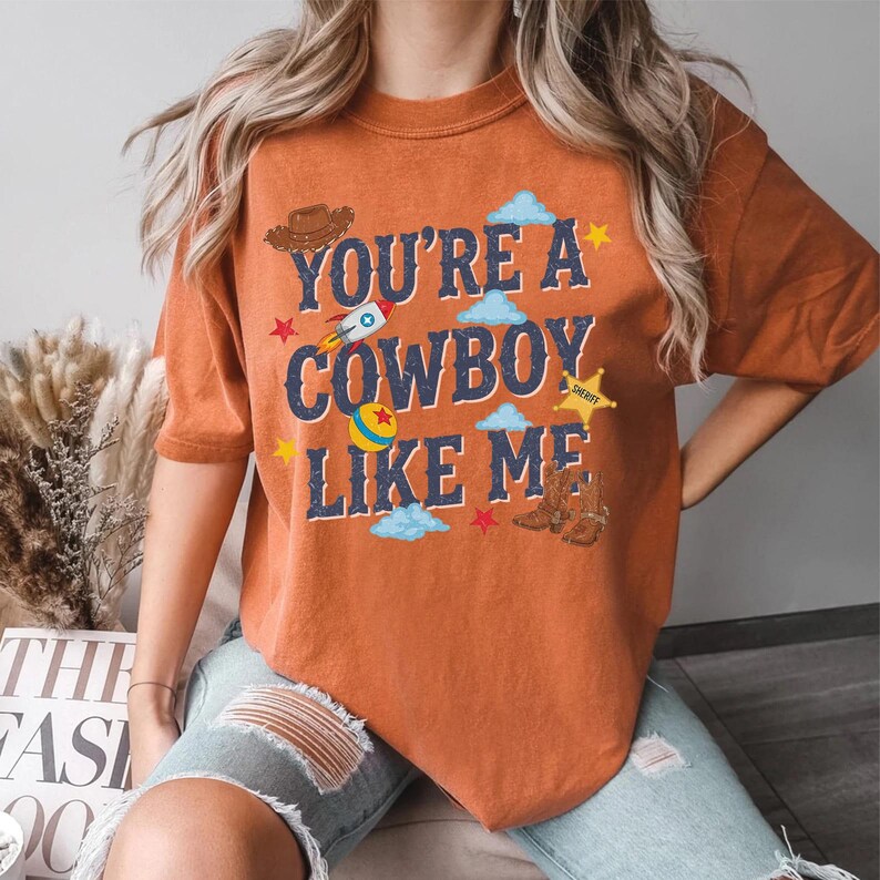 May include: An orange short-sleeved t-shirt featuring the text "YOU'RE A COWBOY LIKE ME" in dark blue letters. The graphic includes a brown cowboy hat, a rocket ship, a colorful ball, a yellow sheriff star badge, brown boots, and blue clouds.