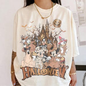 May include: An off-white short-sleeve t-shirt featuring a graphic print of cartoon characters in Halloween costumes standing before a castle. The design includes a full moon, bats, and the word HALLOWEEN in large brown letters at the bottom.