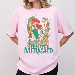 Op de afbeelding: Zachtroze T-shirt met een kleurrijke afbeelding van Ariel uit De Kleine Zeemeermin. Het ontwerp toont Ariel met rood haar en een groene staart, en de tekst "The Little Mermaid" in groen. Het shirt heeft korte mouwen.