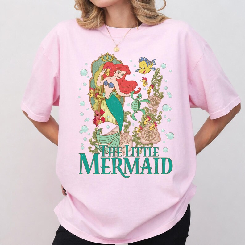 Op de afbeelding: Zachtroze T-shirt met een kleurrijke afbeelding van Ariel uit De Kleine Zeemeermin. Het ontwerp toont Ariel met rood haar en een groene staart, en de tekst "The Little Mermaid" in groen. Het shirt heeft korte mouwen.