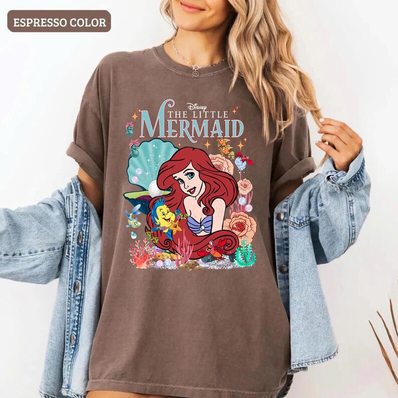 Op de afbeelding: Espresso-kleurige t-shirt met een afbeelding van Ariel uit Disney's De Kleine Zeemeermin. Het ontwerp bevat Ariel, Botje en Sebastiaan, omringd door koraal en bloemen. De woorden "Disney The Little Mermaid" staan boven de afbeelding.