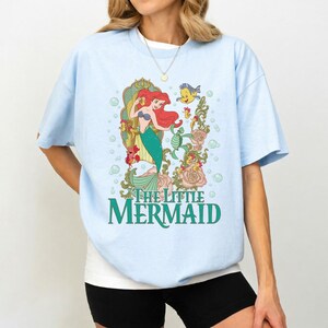 Op de afbeelding: Lichtblauw T-shirt met een kleurrijke afbeelding van Ariel uit De Kleine Zeemeermin. Het ontwerp bevat Ariel, Botje en bloemmotieven, met de tekst "THE LITTLE MERMAID" in groen. Het shirt heeft korte mouwen en een ronde hals.