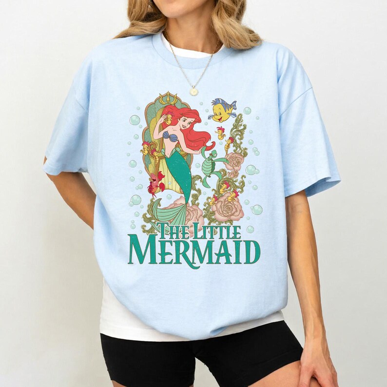 Op de afbeelding: Lichtblauw T-shirt met een kleurrijke afbeelding van Ariel uit De Kleine Zeemeermin. Het ontwerp bevat Ariel, Botje en bloemmotieven, met de tekst "THE LITTLE MERMAID" in groen. Het shirt heeft korte mouwen en een ronde hals.