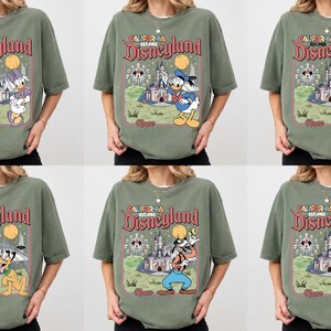 May include: Olive green t-shirts featuring various Disney characters and the Disneyland logo. The graphic includes a castle, Ferris wheel, and the words "California Disneyland Est. 1955". Each shirt showcases a different character: Donald Duck, Mickey Mouse, Pluto, Goofy, and Minnie Mouse.