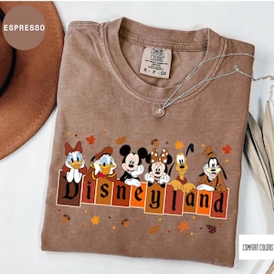 May include: An espresso-colored short-sleeve t-shirt featuring a graphic of Daisy Duck, Donald Duck, Mickey Mouse, Minnie Mouse, Pluto, and Goofy above the text Disneyland. The design includes scattered autumn leaves and acorns on the fabric.
