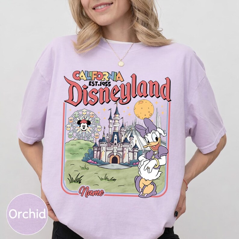 May include: Orchid-colored t-shirt featuring a Disneyland graphic. The design includes the words "California Disneyland EST.1955" in a retro font, along with illustrations of a castle, Ferris wheel, and Daisy Duck.