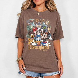 May include: A brown short-sleeve t-shirt featuring a graphic print of Mickey Mouse, Minnie Mouse, Donald Duck, Goofy, and Pluto in front of a castle. The text "Disneyland Est. 1955" is printed in gold lettering below the characters on the front of the shirt.