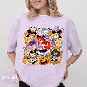 May include: A light purple t-shirt featuring a Halloween-themed graphic. The graphic includes framed illustrations of animated princesses, bats, pumpkins, and the text "Spooky Princess Gathering" and "Happy Halloween."