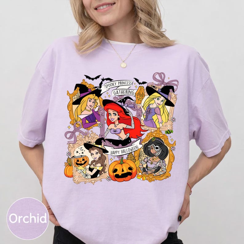May include: A light purple t-shirt featuring a Halloween-themed graphic. The graphic includes framed illustrations of animated princesses, bats, pumpkins, and the text "Spooky Princess Gathering" and "Happy Halloween."