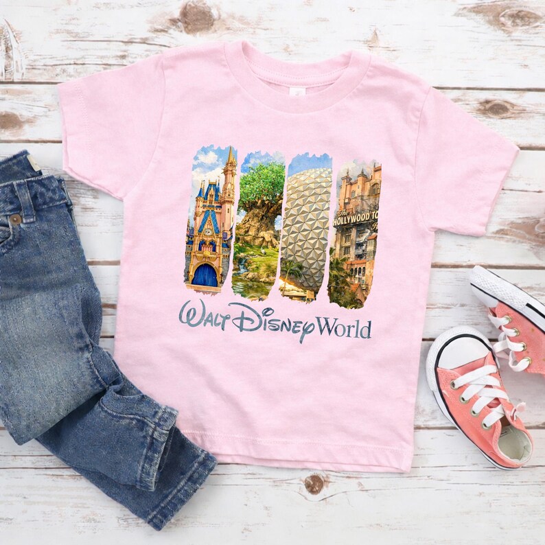Puede incluir: Una camiseta rosa claro con cuatro im&aacute;genes rectangulares verticales de los lugares emblem&aacute;ticos de Walt Disney World. La camiseta tiene las palabras "Walt Disney World" impresas en una fuente cursiva. Un par de vaqueros azules y zapatillas rosas est&aacute;n al lado de la camiseta.