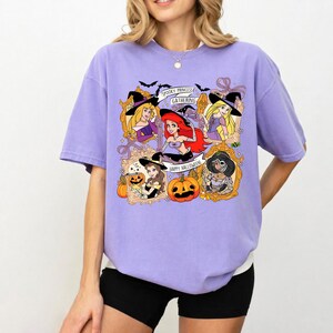 May include: A light purple t-shirt featuring a Halloween-themed graphic. The graphic includes cartoon princesses in witch hats, pumpkins, and the text "Spooky Princess Gathering" and "Happy Halloween".