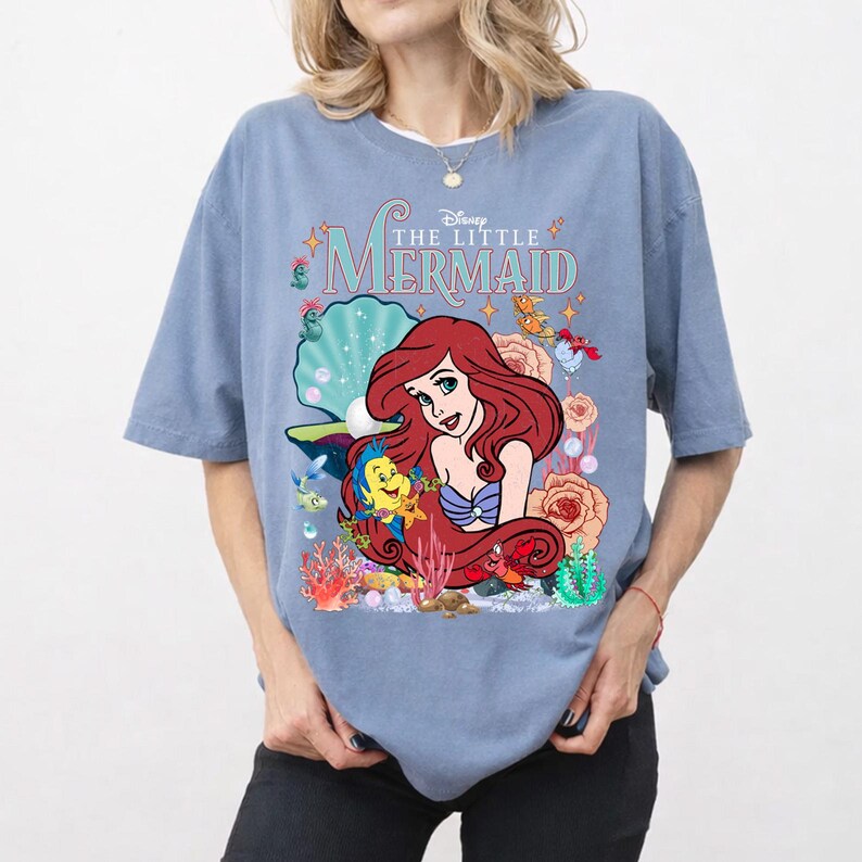 Op de afbeelding: Blauw T-shirt met een afbeelding van Ariel uit Disney's "De Kleine Zeemeermin". Het ontwerp bevat Ariel, Botje, Sebastiaan en de woorden "The Little Mermaid". Het shirt heeft korte mouwen en een ronde hals.