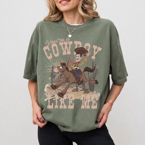 May include: An olive green short-sleeve t-shirt featuring a graphic print of a cartoon cowboy riding a brown horse. The text "You're a COWBOY LIKE ME" is printed in a light pink font above and below the illustration, which includes cacti.