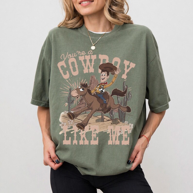May include: An olive green short-sleeve t-shirt featuring a graphic print of a cartoon cowboy riding a brown horse. The text "You're a COWBOY LIKE ME" is printed in a light pink font above and below the illustration, which includes cacti.