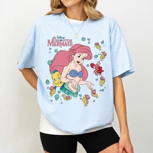 May include: A light blue short-sleeve t-shirt featuring a graphic print of Ariel from The Little Mermaid with Flounder, Sebastian, and seahorses. The design includes the text Disney The Little Mermaid in pink and green with teal bubbles.