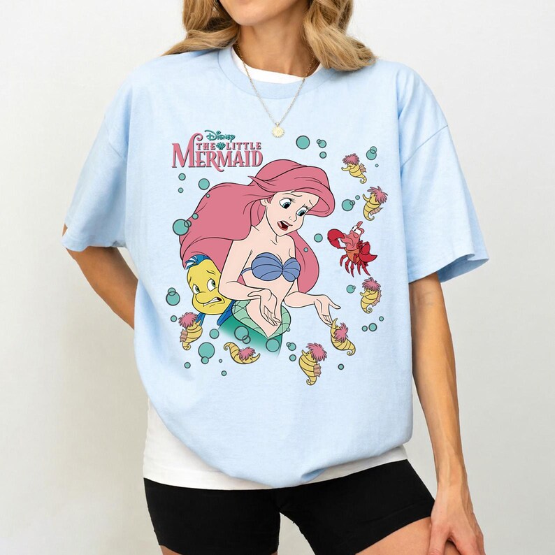 May include: A light blue short-sleeve t-shirt featuring a graphic print of Ariel from The Little Mermaid with Flounder, Sebastian, and seahorses. The design includes the text Disney The Little Mermaid in pink and green with teal bubbles.