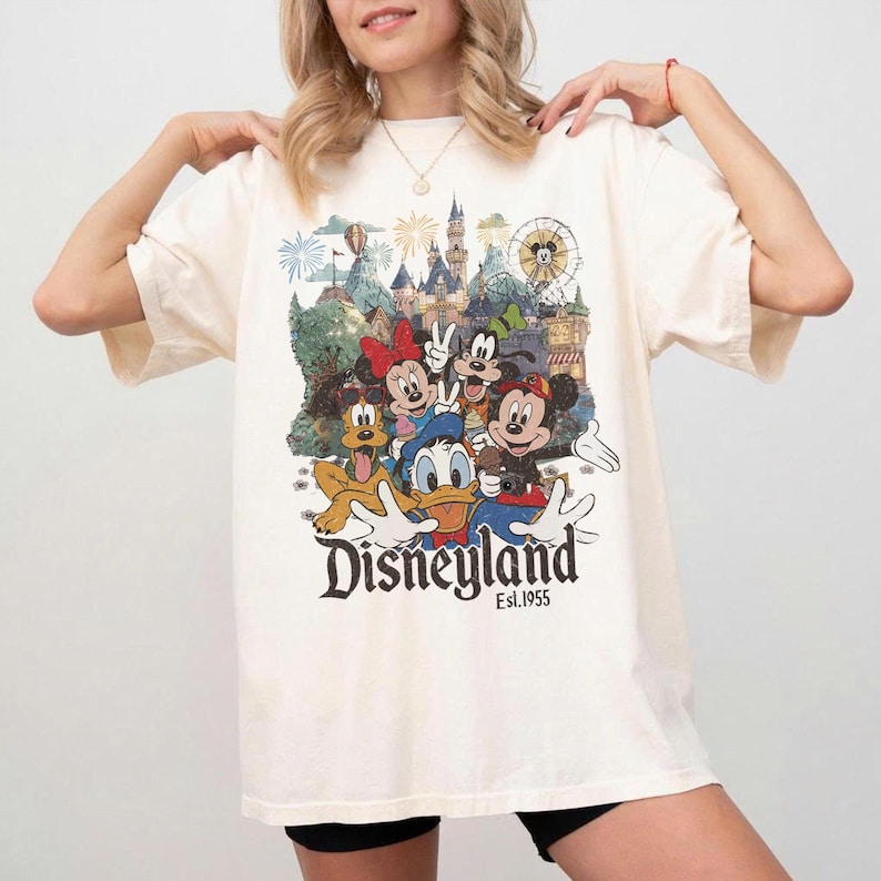 May include: An off-white short-sleeve t-shirt featuring a colorful graphic print of Mickey Mouse, Minnie Mouse, Donald Duck, Goofy, and Pluto in front of a castle. The text Disneyland Est. 1955 is printed in black below the character illustration.